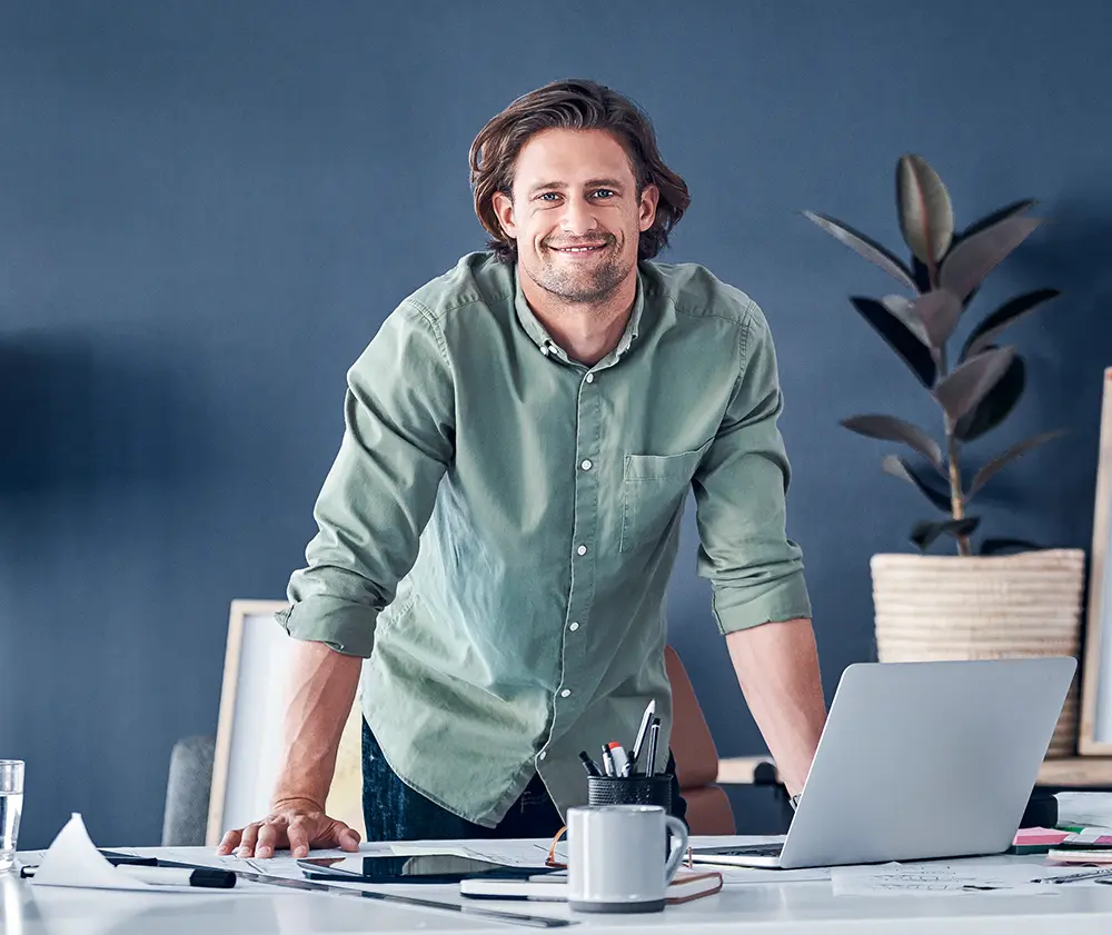 Man in a green shirt standing at a desk with a laptop and papers